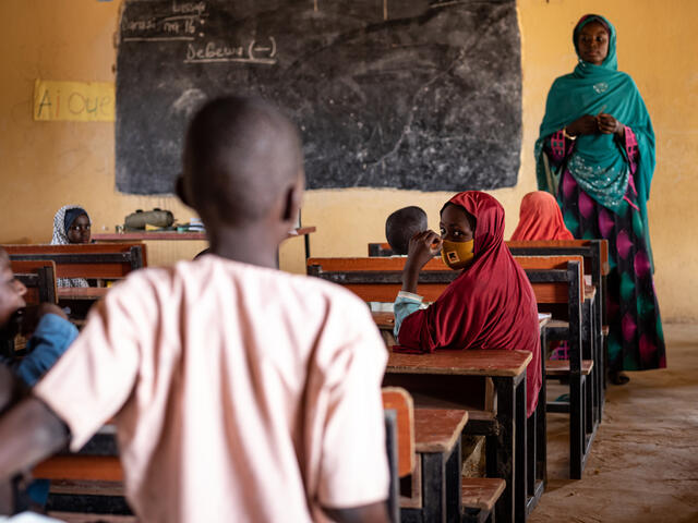 A classroom with the teacher stood at the front of the class next to a blackboard. 