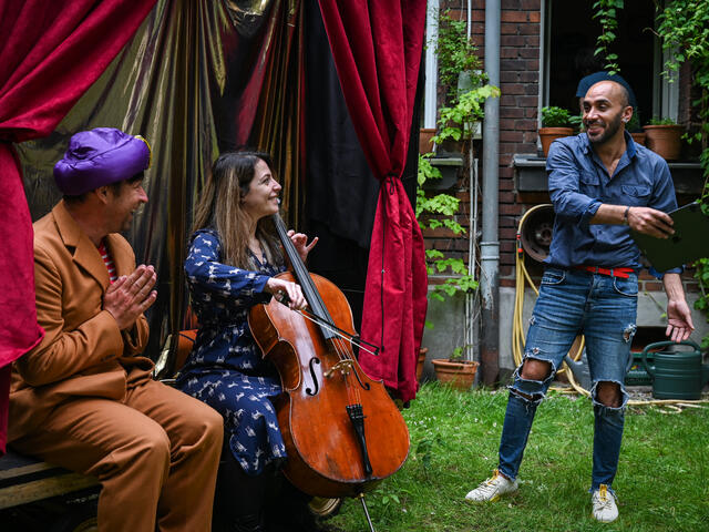 Bashar is standing up and smiling at two performers sat in front of the theatre set. One performer is wearing a turban and the other is playing the Cello. 