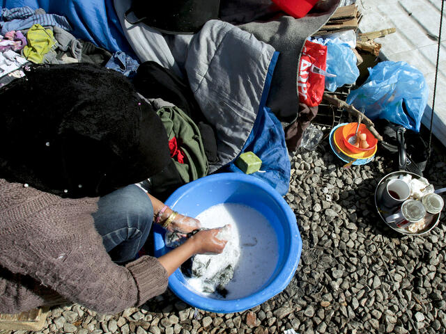 A woman sits on the ground, washing her clothes in soapy water in a plastic basin.