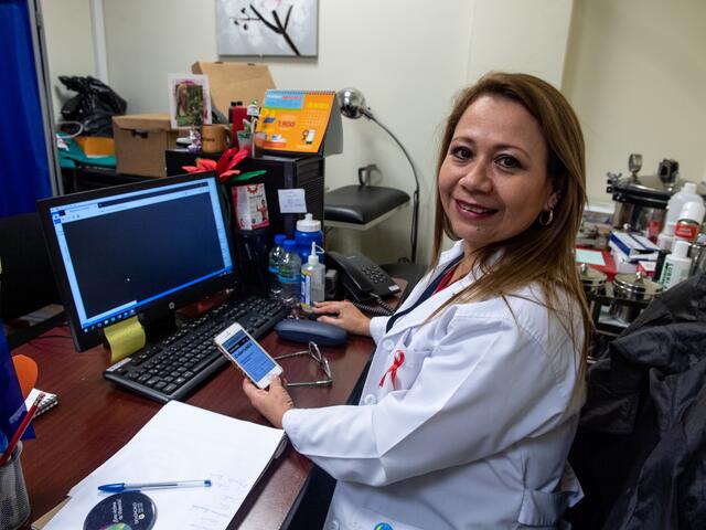 A doctor at her desk in San Salvador holds a phone displaying CuentaNos.org on its screen.