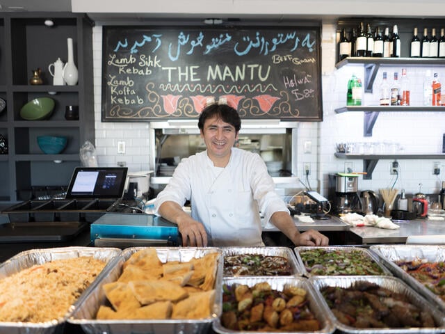 A chef from Afghanistan stands behind the counter of his restaurant with trays of Afghan food in front of him.