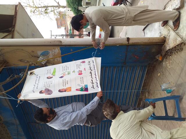 Two men wearing masks stand on either side of a poster with COVID-19 information. 