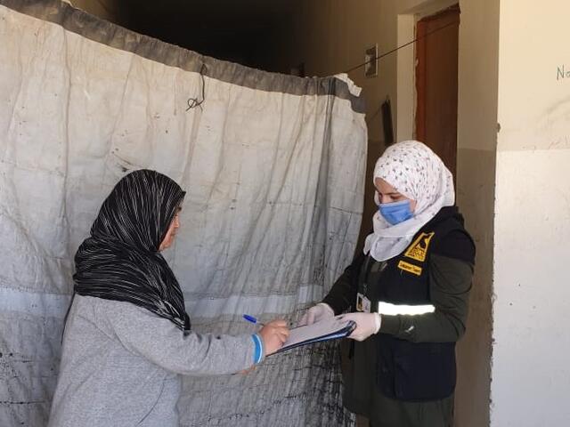 A woman in an IRC vest holds a clipboard while talking to an older woman and distributing cash assistance. 