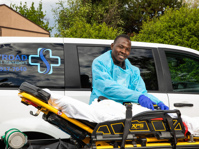 Jonathan Amissa, wearing scrubs, stands in front of a van with his hands on a stretcher. The van has a logo for Skyroad Medical Transport, his medical transportation business. 