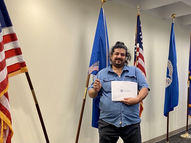 Former refugee, Noor, holding his United States Citizenship Certificate with United States flag and Utah flag in the background.