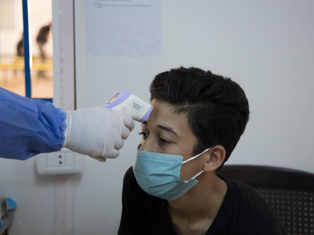 Wearing a mask, Muhammad sits in a clinic while a doctor wearing full PPE takes his temperature. 