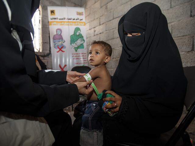 An IRC health worker in Yemen measures a toddler's upper arm for signs of malnutrition as he sits on his mother's lap.