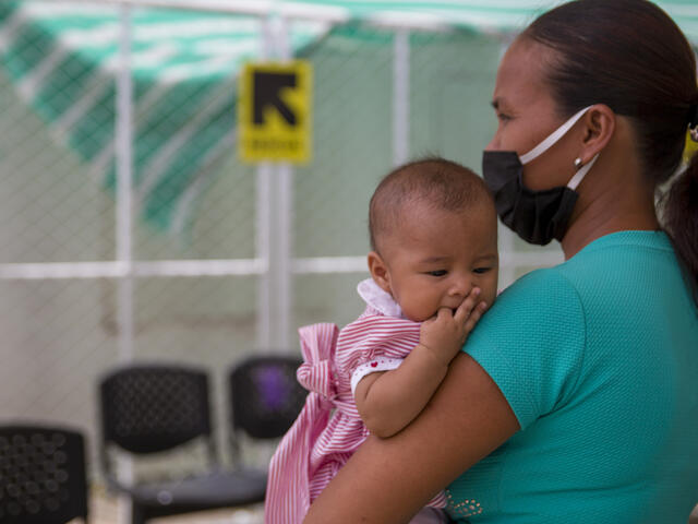 A mother wearing a face mask holds her baby daughter in the socially distanced waiting area of an IRC clinic on the Colombia-Venezuala border during the COVID-19 pandemic.