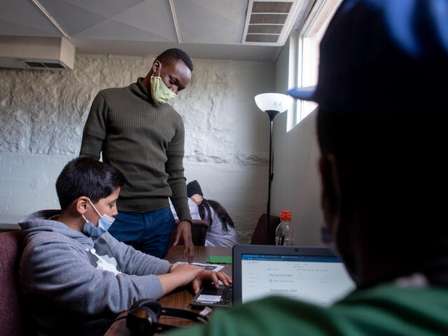 Fredrick stands next to a student at an after-school program. Both are wearing masks and the student is on his computer while Fredrick looks over his shoulder. 