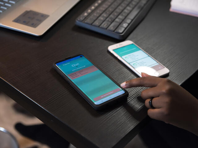 Diana points to her app open on a smart phone on a brown table. 