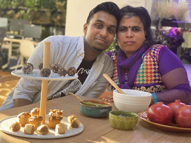 Sitting at a table with an array of food in front of them, Shanthini and Sarujen lean toward one another and look at the camera. 