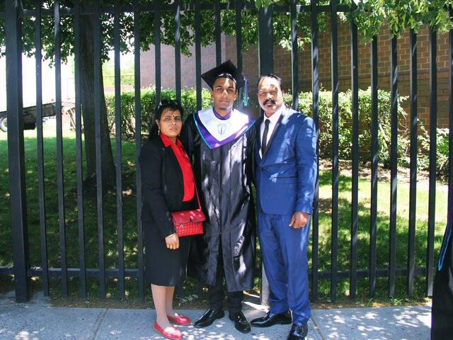 Wearing a graduation cap and gown, Sarujen stands in front of a fence with his parents on either side of him. 