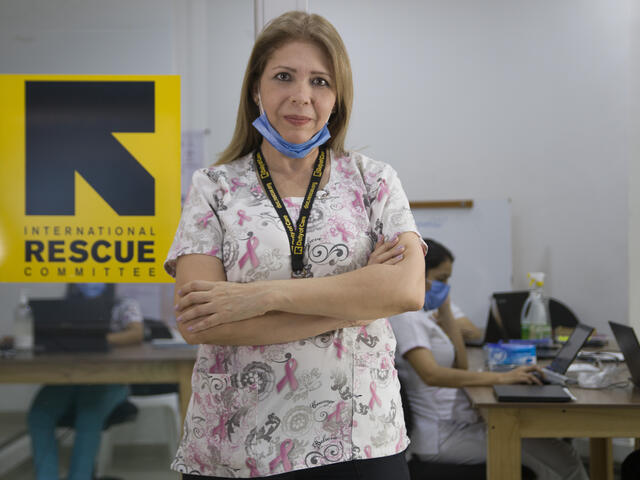 Dr. Edna Patricia Gomez stands in front of an IRC logo in a clinic wearing her medical scrubs and a mask around her chin. 