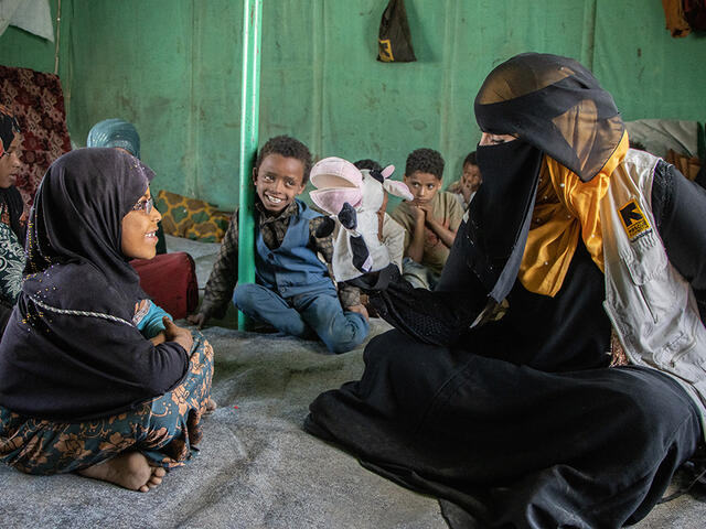 A group of young children sit while watching a puppet show. One girl interacts with the volunteer talking to her with a cow puppet