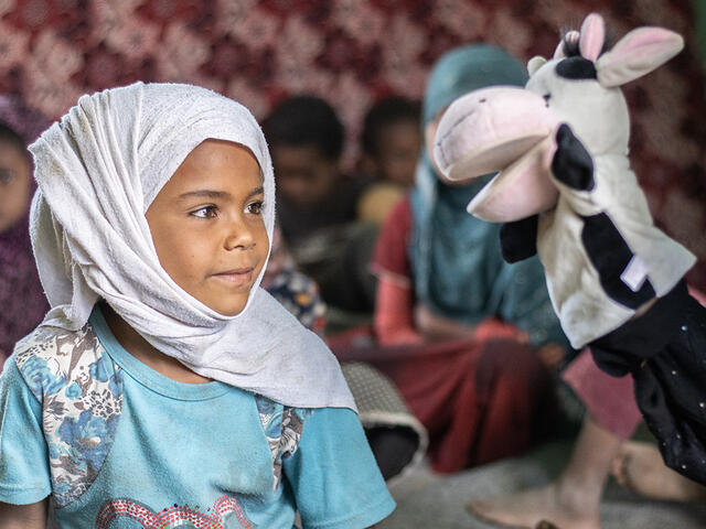 In Al-Manshar camp, a litle girl sits and looks at a cow puppet that a volunteer is using to teach her about COVID-19 prevention.