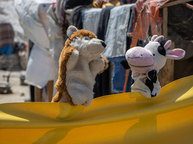 Using a yellow tarp as their "stage," a cow and fox puppet have a "conversation" during a puppet show on COVID-19 prevention 