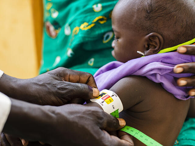 A mom holds a small child on her lap while we see the hands of a community health worker holds a MUAC (middle upper arm circumfrence) band around his arm. 