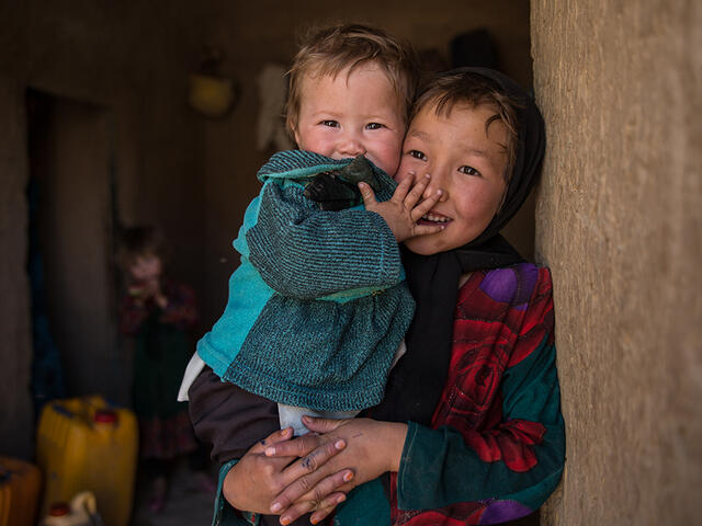 A girl holds her younger sister and looks at the camera, both are smiling and laughing. 