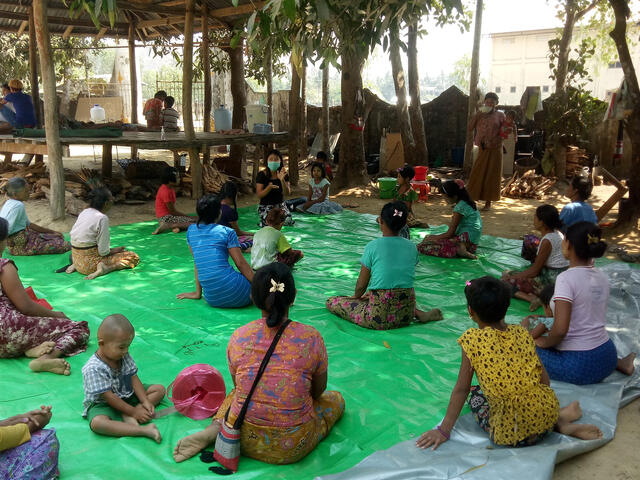 People listen to an IRC health worker provide health information. They are sitting outdoors socially distanced on a mat as a COVID-19 precaution.