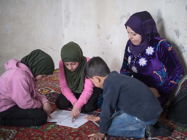 Reema sits on the ground with  her twins Shurouq and Shirina and their brother Eissa. Their children are looking at schoolwork while Reema looks over their shoulders. 