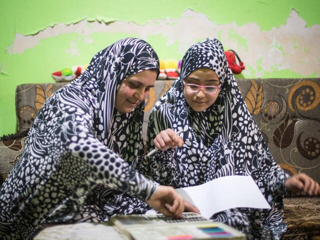 Wearing matching outfits, Ruba and Salam sit on a couch while Ruba helps her Salam with her homework. 
