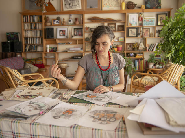 Diala, sitting at her desk holding a pencil, reviews preliminary sketches of her "Refugees are courageous" illustration. 