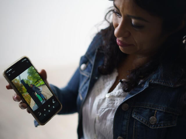 Wearing a jean jacket, Rosa holds out her iPhone. There is a picture of her daughter on the screen. 