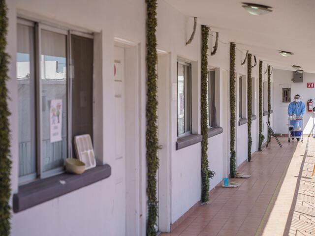 A health care worker in full PPE pushes a cart down an outdoor walkway in front of doors to hotel rooms. 