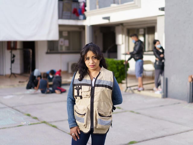 Rosa, wearing a brown vest with large pockets, stands in front of the triage hotel she helps manage in Ciudad Juárez, Mexico. In the background, children draw with chalk on the ground with their parents while other adults converse. 
