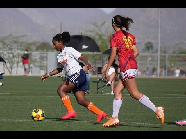 Kezia kicks a soccer ball while another player approaches her 