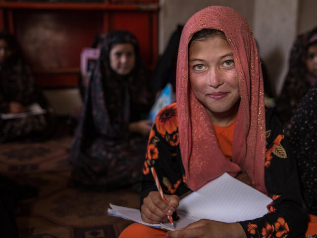 A young girl sits with a notebook on her lap ready to write while looking up at a teacher. There are other young girls sitting behind her. 