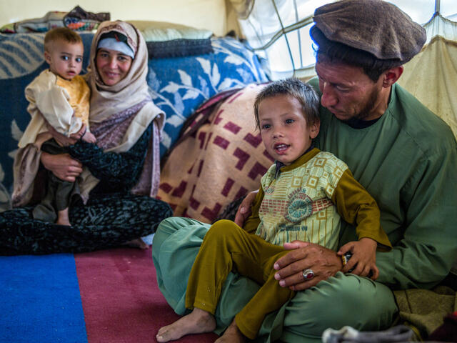 A family of four sit on rugs on the ground: a young boy sits on the lap of his father and a baby sits on the lap of her mother.