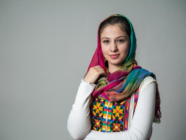 A portrait of Muska Haseeb on a grey background. She is a young woman in her early 20s wearing brightly colored clothes. 