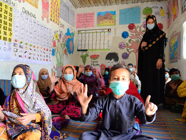 Children sit on the floor and a female teacher stands in a classroom, all are wearing masks and the kids are clapping. 