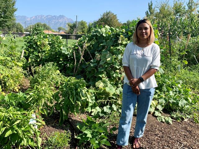 Gardener Menuka stands in front of her garden featuring various green plants.