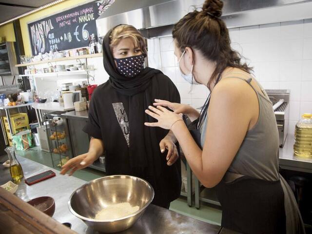 In a kitchen, two women, both masked, talk to one another as they mix flour and spices in a large bowl. 