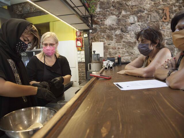 In a kitchen, Hafea stands next to one woman and shows her a technique while two additional woman sit on the other side of a counter watching. 