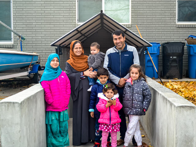A family of two adults and five children smile in front of the entrance to their new home.