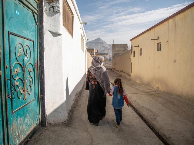 With their backs to the camera, Noor and her daughter walk between two buildings holding hands. There are mountains in the distance. 