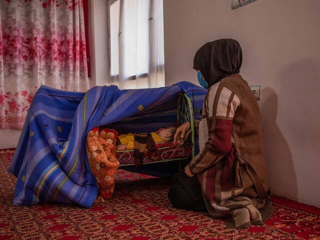 Zulaykha kneels on the ground next to her one-year-old son, who is sleeping bassinet on the ground shielded by blankets.