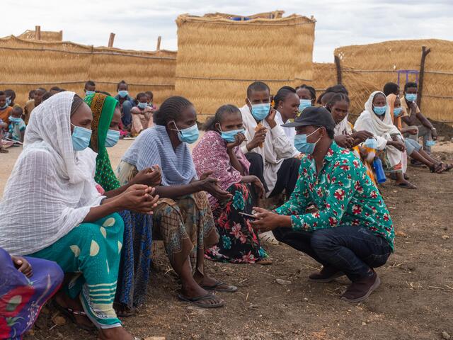 People in Tunaydbah camp in Sudan sit on benches while one man kneels in front of them. 