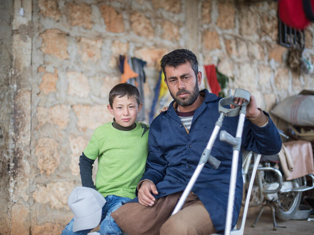 A man missing one of his legs sits holding crutches while his son sits next to him.