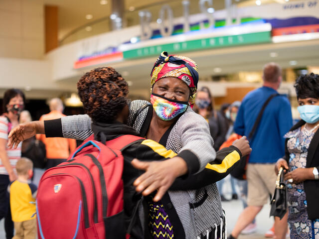 In an airport, a middle aged woman hugs a teenager with a back pack. She is wearing a mask but it is clear she is smiling and emotional.  