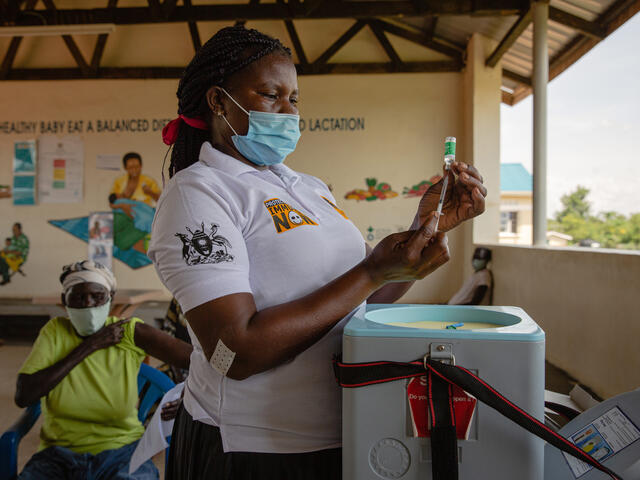 In the foreground, a health care worker prepares a syringe while a patient in the background rolls up her sleeve to receive the vaccine. 