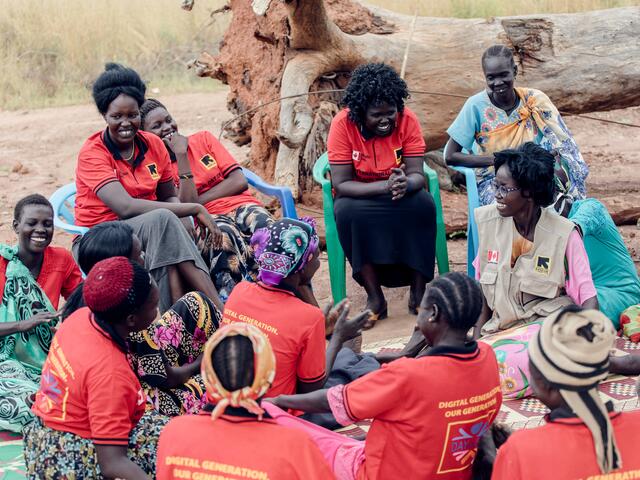 A group of women wearing matching red shirts with IRC logos sit in a circle outside, some on chairs and some on a blanket. They are smiling and talking.  