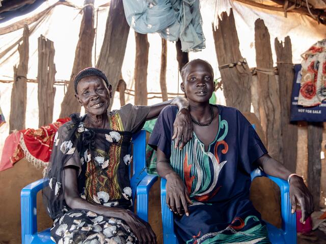 Two women sit on chairs, with one resting her arm on the other's shoulder. Both are smiling.