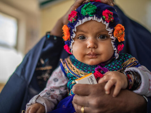 A close up a five-month-old baby sitting on her mothers lap and wearing a brightly colored knit hat.