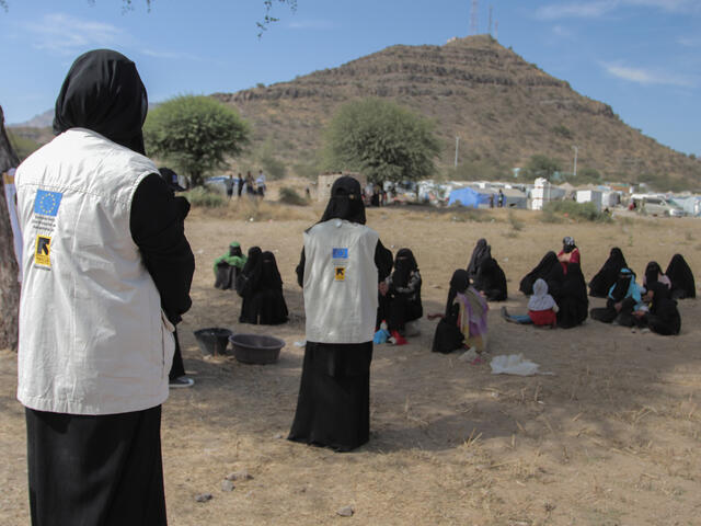 Two women stand in IRC-branded vests teaching to other women sitting on the ground. They are outside with a mountain in the background. 