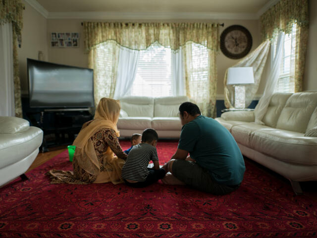 With their back to the camera, a husband and wife sit on their living room floor with their two children. The room also has a tv and white sofa. 