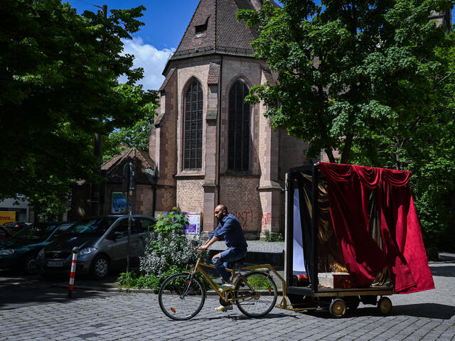 On a three-lined street, Bashar is sitting on a bicycle that has a stage with a red curtain attached to it.